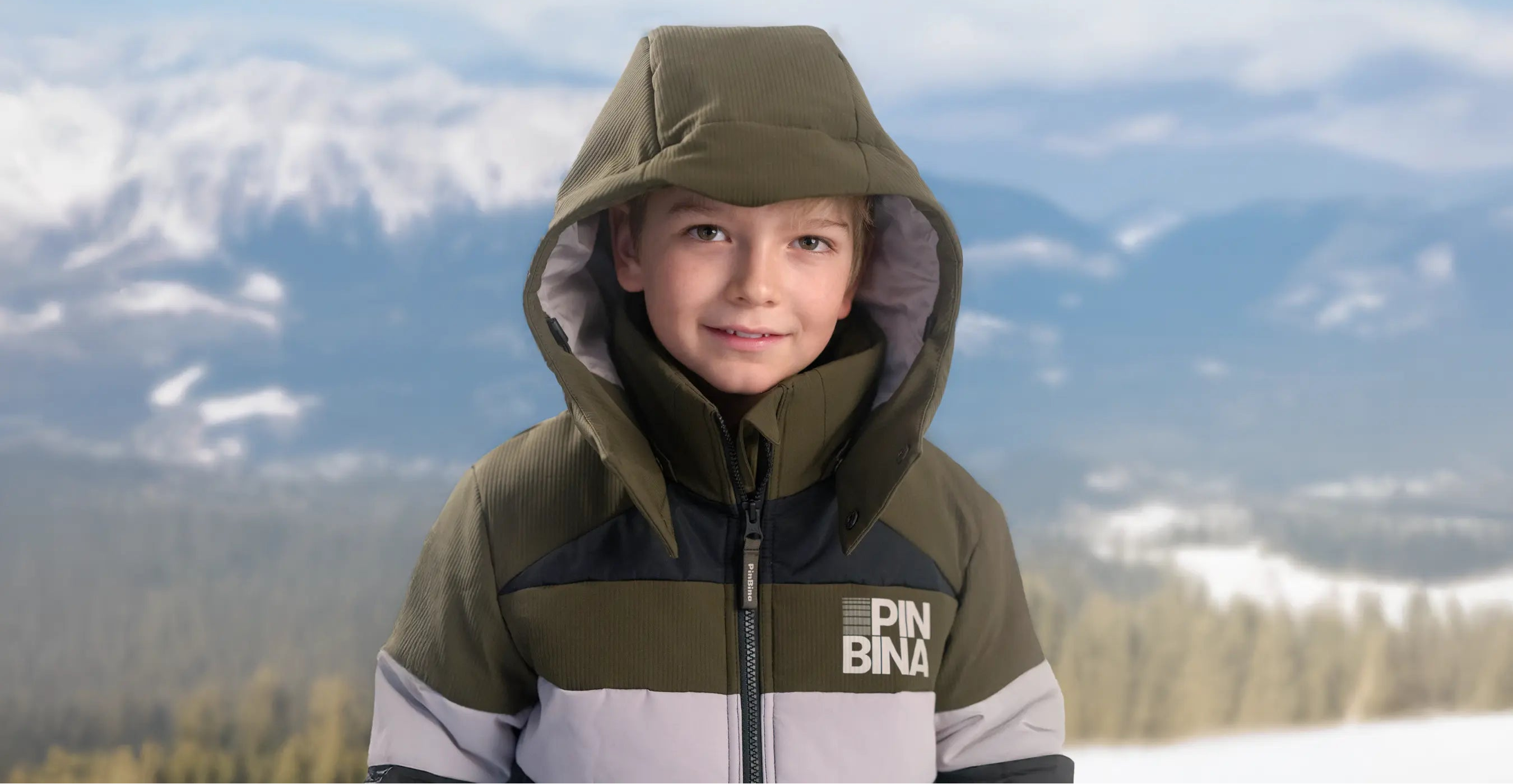 Child wearing a green and gray jacket with 'PIN BINA' logo against a mountainous landscape.